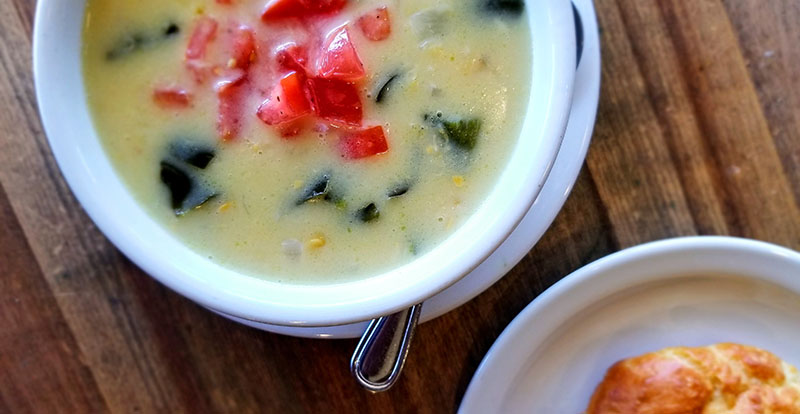 Cup of poblano corn chowder with tomato garnish and buttermilk biscuit.